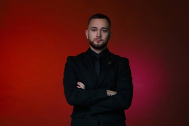 Kırmızı arka planda siyah klasik takım elbiseli yakışıklı bir adam kollarını kavuşturmuş duruyor..