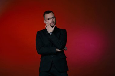 Siyah klasik takım elbiseli, kırmızı arka planda sakallı yakışıklı bir adam ellerini yüzüne doğru tutuyor..