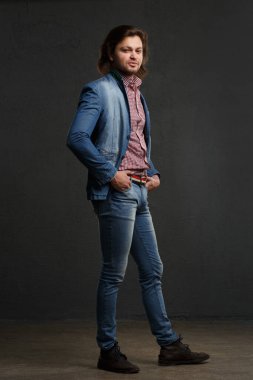 An attractive white man in jeans stands in full growth against a dark background.
