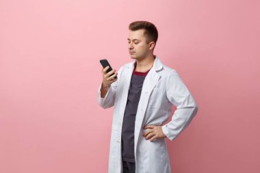 Young doctor in a white coat carefully looks at the smartphone screen isolated on a pink background.