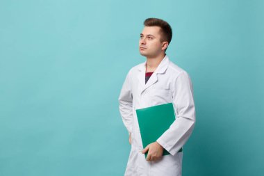Young handsome doctor in a white coat holding a green folder isolated on a blue background.