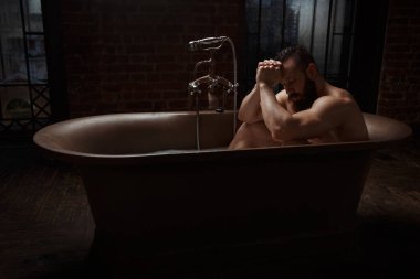 Atletik sakallı adam karanlık bir çatı katında şık bir banyoda dinleniyor..