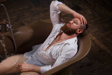 Atletik sakallı bir adam karanlık bir iç mekanda lüks bir banyoda dinleniyor..