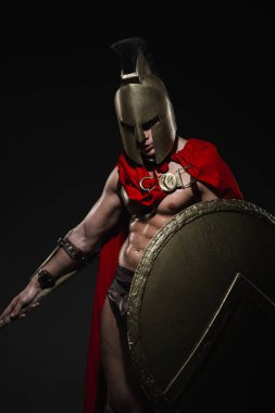 Siyah arka planda kırmızı pelerinli ve kasklı Romalı bir savaşçı kostümü giymiş genç atletik seksi bir adam..