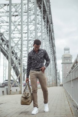 Elinde büyük bir çantayla genç ve çekici bir siyah adam şehirde köprüden geçiyor..