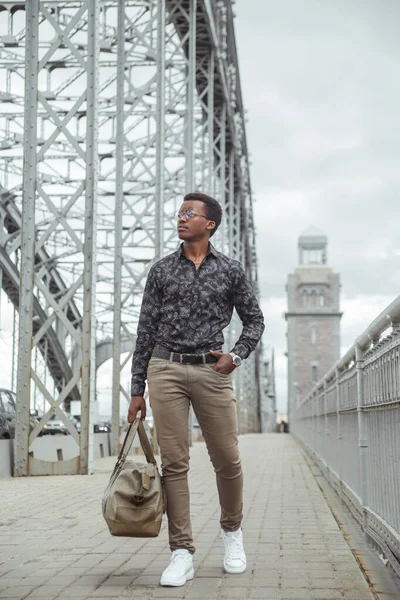 Elinde büyük bir çantayla genç ve çekici bir siyah adam şehirde köprüden geçiyor..