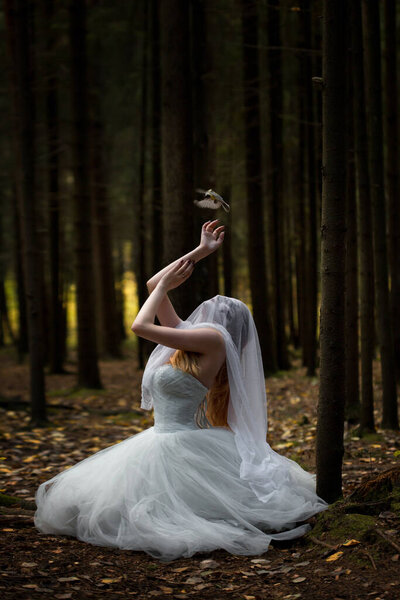A mysterious woman bride with a veiled face in a gloomy forest in a prayer position with a bird crawling into her hand.