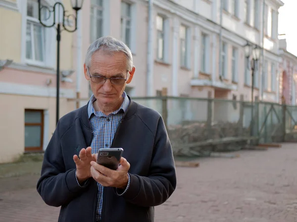 Elderly person in the glasses is holding a mobile phone in his hands. Senior man is texting on smartphone. Old man is using cellphone