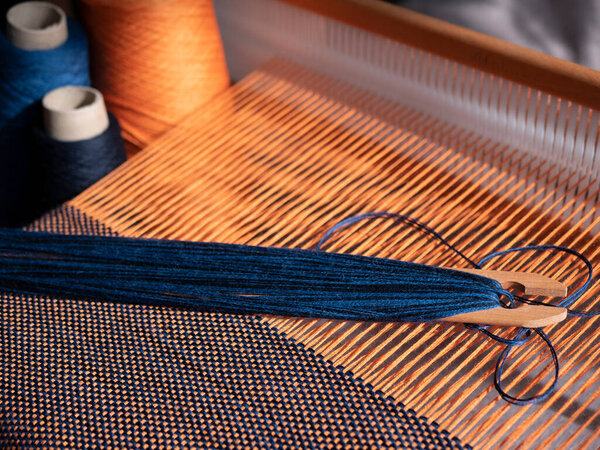 Creation of the blue and orange textile using weaving loom. Wooden loom, shuttle and cones with yarn. Clothing fabricating with a handloom