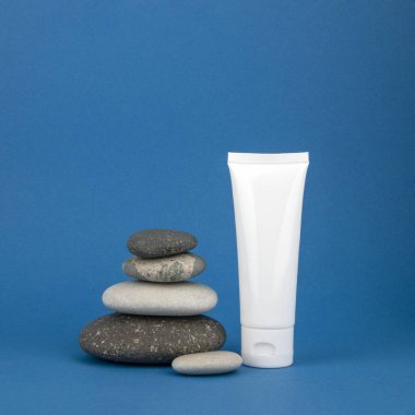 Mavi arkaplandaki krema için beyaz tüp. Yuvarlak doğal taşlar piramidi, kopyalama alanı. Kaplıca bakımı ve güzellik kavramı