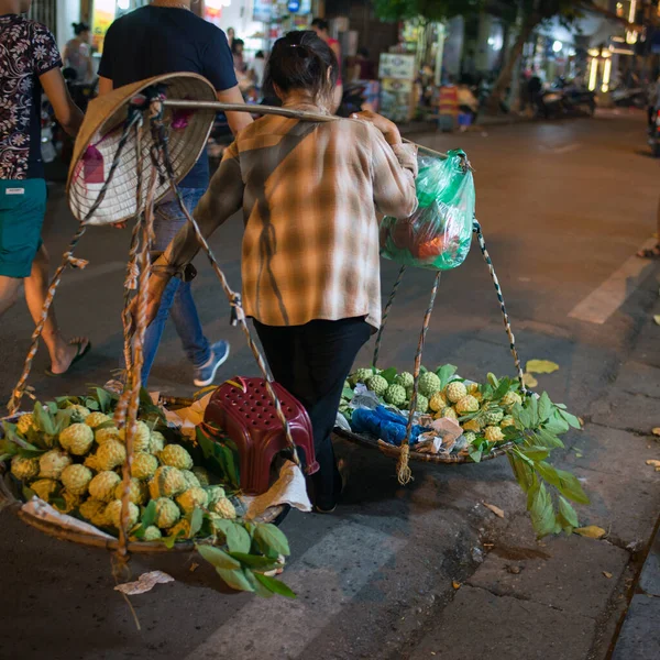 Street vendor starting her work. Backview of a woman carrying a shoulder pole with fresh fruits. Straw conical hat. City lights on. Hanoi, Vietnam. Asia.