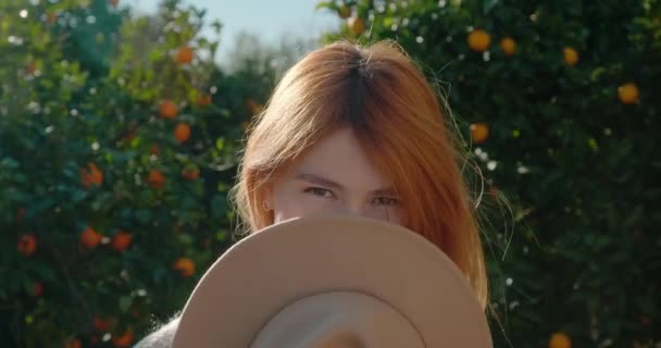 Young woman covers her face with hat and poses, standing against orange trees — Vídeo de Stock