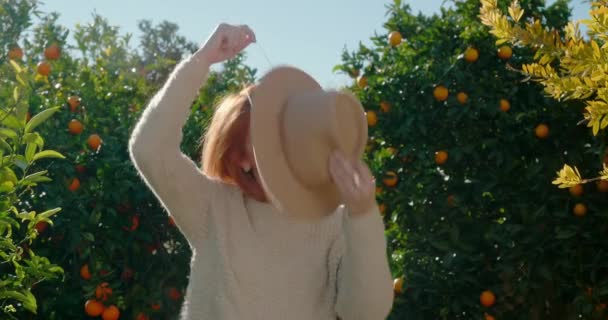 Young woman puts on a stylish hat and poses, standing against orange trees — Vídeo de Stock