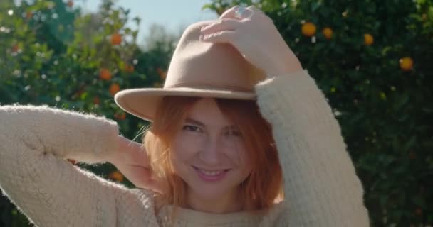 Young woman puts on a stylish hat and smile, standing against orange trees — стоковое видео