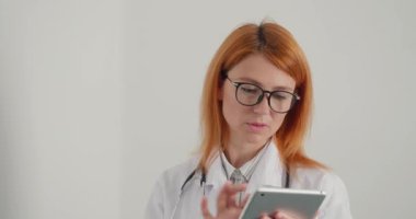 Gözlüklü kızıl saçlı kadın doktor tablet üzerinde çalışıyor. Doktor hastalara çevrimiçi danışmanlık yapıyor. Ciddi bayanın muayenehanede her zamanki çalışma günü.