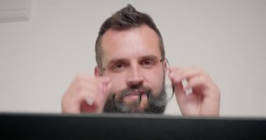 Brunet sakallı adam kameraya bakarken gözlüğü çıkartıyor. Ofiste bej duvarın karşısında oturan çalışanın portresi dizüstü bilgisayarla yakından görülüyor.