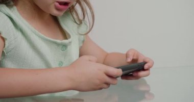 Ciddi anaokulu kızı, mutfaktaki cam masada otururken akıllı telefondan ilginç oyunlar oynuyor. Çocuk beyaz arkaplandaki yakın plan için aygıt kullanıyor