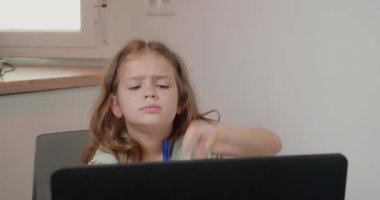 Sıkılmış kız masada oturup dizüstü bilgisayarla öğretmenin açıklamasını dinliyor, baş parmağını kaldırıyor. Okul çocuğu çevrimiçi ders sırasında elini yüzüne koyar ve uykulu hisseder.