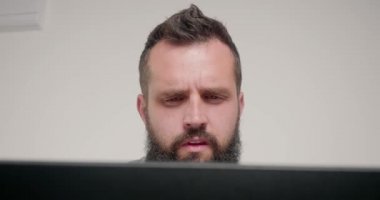 Konsantre çalışan, dizüstü bilgisayara bakarak gözlük takar. Sakallı adam, ofiste bej rengi duvara karşı otururken gözleri gerginlikten korur.