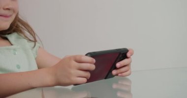 Şirin küçük kız ağzıyla ilginç bir oyun oynuyor. Çocuk cam masada oturuyor ve elinde beyaz arka planda akıllı telefon var.