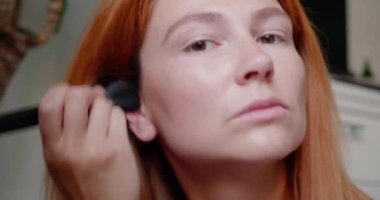 Aynadaki kızıl saçlı kadının yansıması her zamanki sabah rutinini yapıyor. Kadın yüzüne pudra sürerek makyaj yapıyor. Kadın yakın çekim için hazırlanıyor.