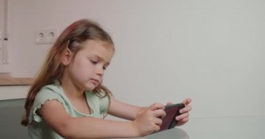 Uzun sarı saçlı küçük kız modern akıllı telefondan eğlenceli oyunlar oynamaktan hoşlanıyor. Odaklanmış çocuk mutfak masasında yakın plan cihazla oturuyor.