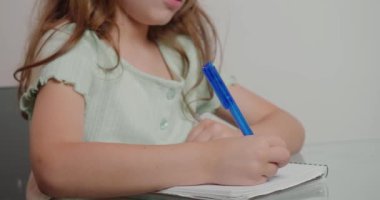 Liseli kız, egzersiz kitabında kalemle yazma işi yapıyor. Uzun saçlı küçük kız cam masada oturur ve ev ödevlerini yakın plan yapar.