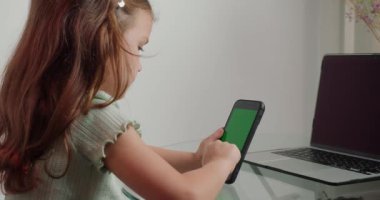 Küçük kız bilgisayarın yanındaki cam masada oturan kromakey ekranlı akıllı telefondan oyun oynuyor. Liseli kız çevrimiçi dersten sonra ara veriyor.