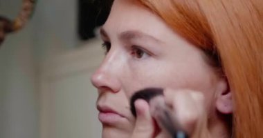 Kızıl saçlı kadın yüzüne pudra sürerek makyaj yapıyor. Bayan aynaya bakar ve işe hazırlanır. Güzel bir kadının her zamanki sabah rutini vardır.