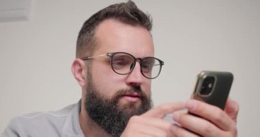 Sakallı adam iş yerindeki sosyal ağ sayfalarını kaydırmak için akıllı telefon kullanıyor. Genç çalışan, bej arka planda gözleri ovalamak için gözlüklerini çıkarıyor.