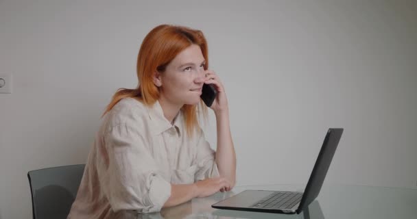 红头发的女人打完电话 继续在笔记本电脑上工作 忙碌的经理坐在办公室的玻璃桌旁 输入新项目的报告 — 图库视频影像