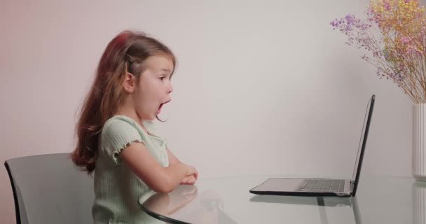 小さな女の子はラップトップ経由でオンラインクラスで退屈します 可愛いです女子高生Yawns見ますのスクリーンのデバイス座っていますデスク近くの花瓶と花 — ストック動画