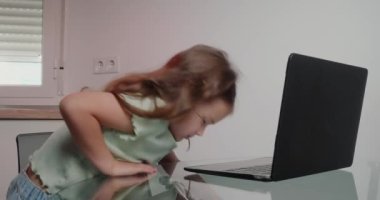 Küçük kız, internetten ders almak için cam masada dizüstü bilgisayarını açıyor. Uzun saçlı tatlı kız evde modern aletler kullanıyor.