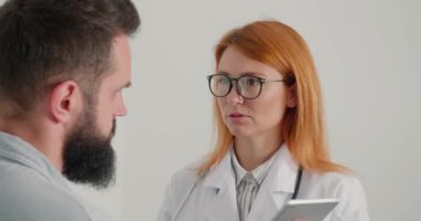 Elinde tabletle gezen kadın doktor, muayenedeki erkek hasta sırasında hapşırıyor. Palto giyen genel pratisyen kadın sakallı adama tedavi prosedürünü açıklıyor.