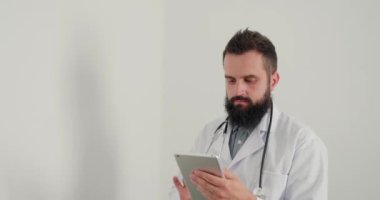 Erkek doktor klinikteki tablette internette sörf yapıyor. Sakallı pratisyen doktor hastalara danışmak için bilgi arıyor ve kameraya bakıp gülümsüyor.
