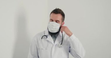 Erkek doktor tıbbi maske takıp kameraya bakıp gülümsüyor. Steteskoplu sakallı pratisyen hekim klinikteki salgın sırasında maskeyle çalışır.. 