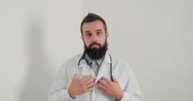 Steteskoplu doktor gülümseyip kameraya bakarak tavsiye veriyor. Doktor hastalara online tavsiye veriyor. Beyaz önlüklü sakallı terapistin klinikte her zamanki işi var.. 