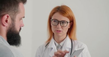 Tabletli kadın doktor, ofisindeki erkek hastaya danışıyor. Palto giyen genel pratisyen kadın sakallı adama tedavi prosedürünü açıklıyor.