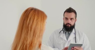Erkek pratisyen doktor klinikteki olgun kızıl saçlı kadına muayene sonuçlarını veriyor. Sakallı doktor randevuda güçlü tavsiyelerde bulunur.