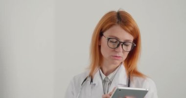 Kızıl saçlı kadın doktor tablet üzerinde çalışıyor. Tıp çalışanı hastalara çevrimiçi danışmanlık yapıyor. Odaklanmış kadın klinikteki uzaklığa bakar ve düşünür.