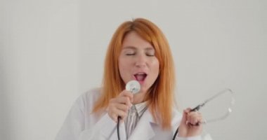 Kızıl saçlı kadın doktor klinikte eğleniyor. Mikrofon olarak steteskobu olan genç pratisyen kadın şarkı söyleyip dans etmeyi sever.