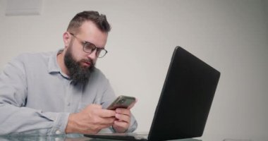 Sakallı adam dizüstü bilgisayarda çalışmak yerine akıllı telefondan sosyal ağlara bakıyor. Gözlüklü eleman ofiste masa başında oturuyor ve aletin dikkatini dağıtıyor.