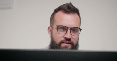 Gözlüklü neşeli sakallı adam ofiste bilgisayarın yanında oturuyor. Profesyonel programcı bej duvar kapanışına karşı yeni proje üzerinde çalışıyor