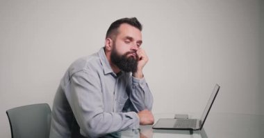 Düşünceli adam ofisinde oturmuş yeni proje için fikir üretiyor. Klasik gömlek giyen genç sakallı bir çalışan dizüstü bilgisayarla çalışıyor.