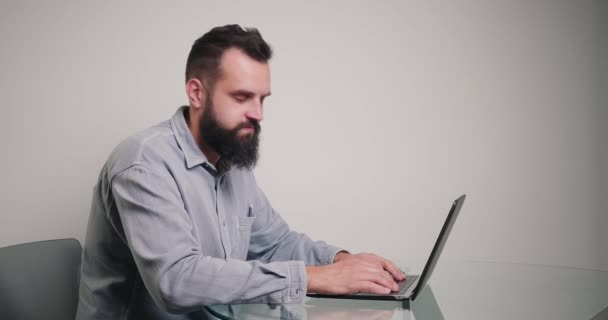 若いコピーライターは カメラへのラップトップの回転の記事をタイプする 古典的なシャツのひげを生やしたブルネットの男は 自宅のオフィスからオンラインで作業机に座っている — ストック動画