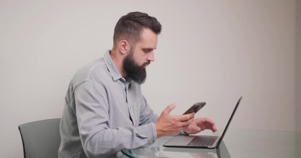 Vousatý Muž Píše Detaily Notebooku Dívá Displej Chytrého Telefonu Ruce — Stock video