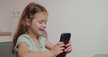 Uzun sarı saçlı küçük kız modern akıllı telefondan eğlenceli oyunlar oynamaktan hoşlanıyor. Odaklanmış çocuk mutfak masasında yakın plan cihazla oturuyor.