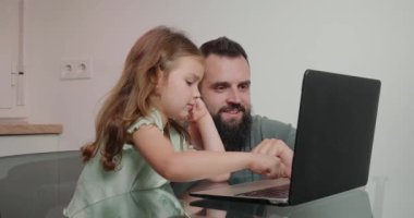 Babası, cam masada oturan küçük kıza dizüstü bilgisayarda ödev yapmasına yardım ediyor. Sakallı adam duygusal olarak kıza elektronik aletin nasıl kullanılacağını anlatıyor.