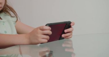 Şirin küçük kız ağzıyla ilginç bir oyun oynuyor. Çocuk cam masada oturuyor ve elinde beyaz arka planda akıllı telefon var.