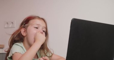 Şirin kız, modern bir bilgisayarla arkadaşlarıyla konuşurken burnunu karıştırıyor. Uzun sarı saçlı mutlu çocuk sandalyeye oturup yakın çekime bakıyor.